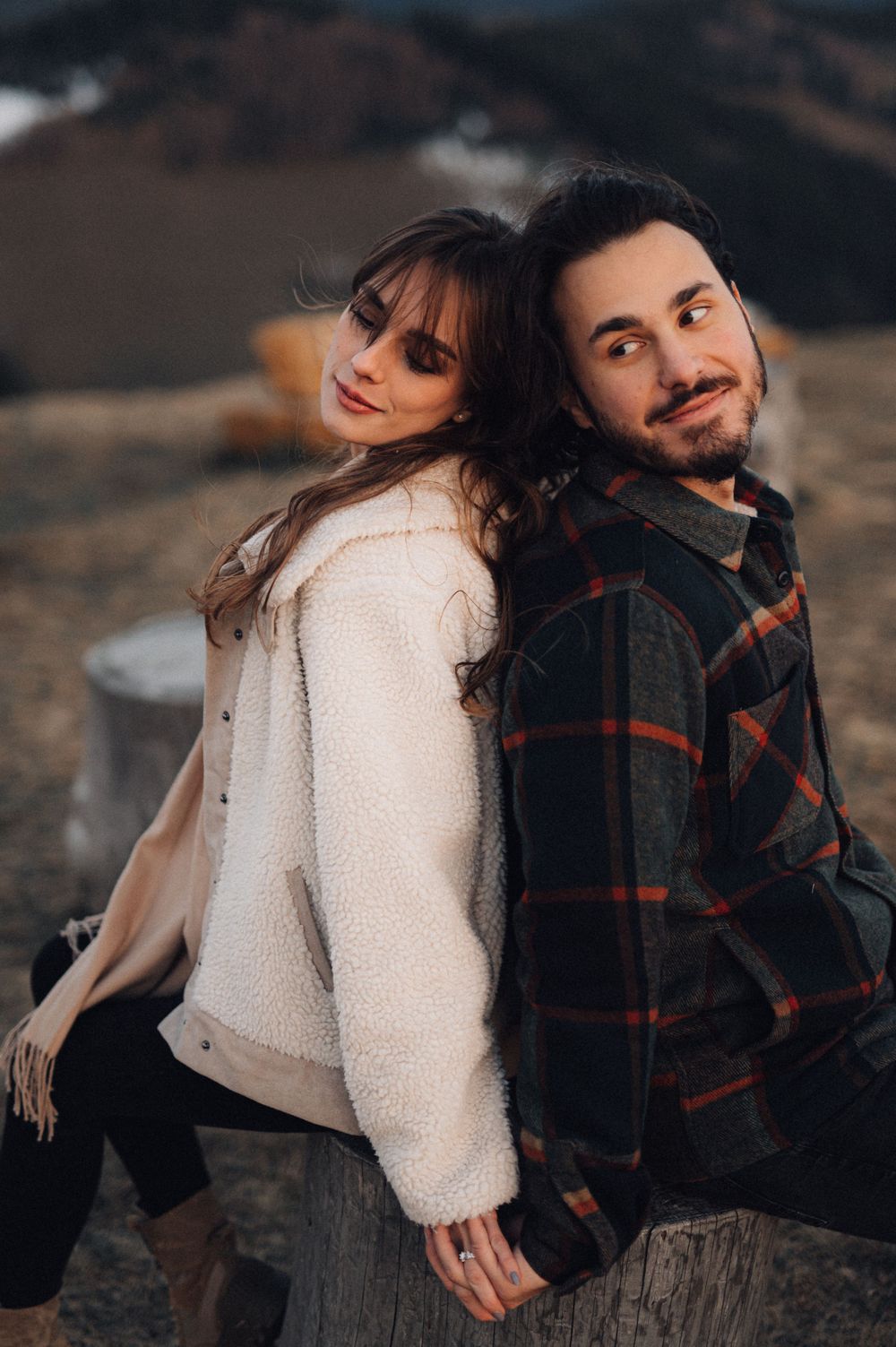 A couple poses together on a wooden stump with mountains visible in the background.