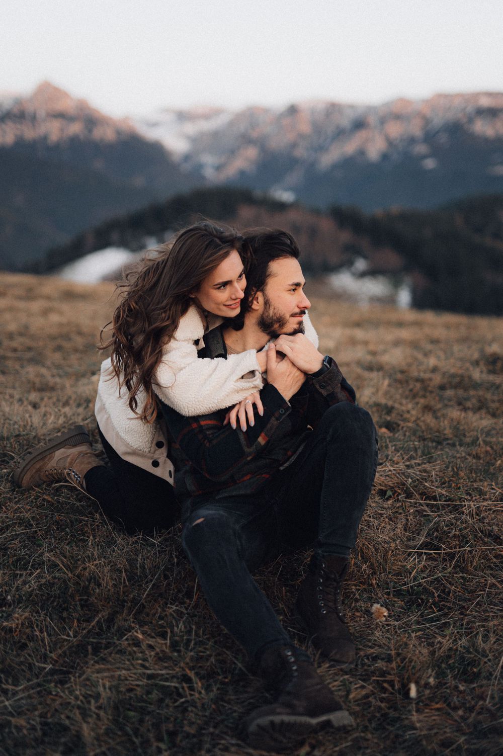 A cozy winter photoshoot features a couple embracing on a hillside with mountains in view.