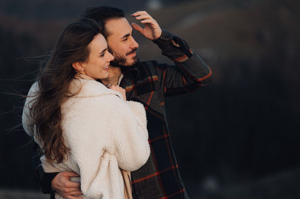 A romantic couple embraces while laughing together outdoors at dusk wearing cozy winter clothing.