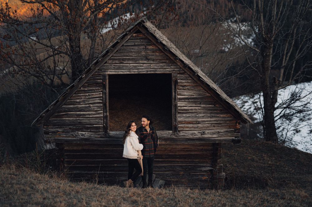 Romantic couple stands in front of weathered wooden barn at dusk.