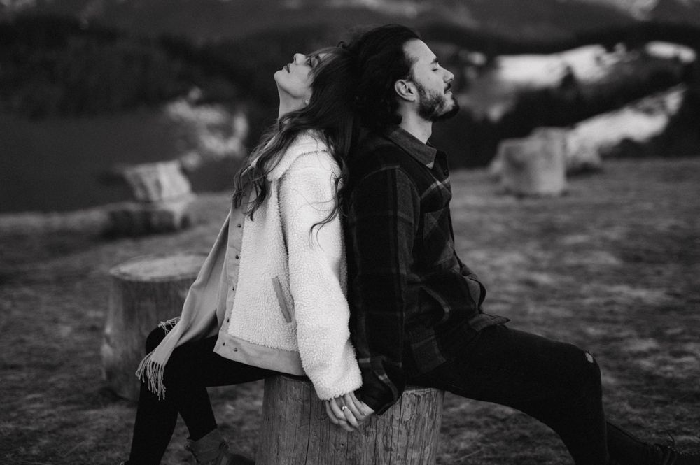A black and white photo of two people sitting back-to-back on a wooden stump in the mountains.