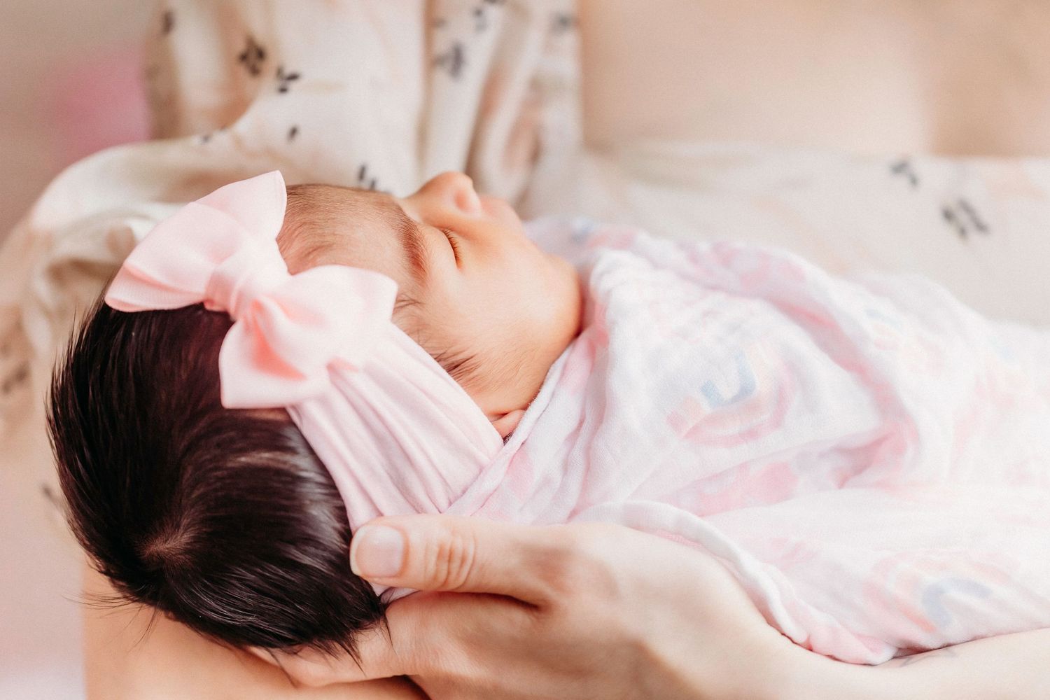 A sleeping newborn wearing a pink bow and floral outfit being cradled gently.
