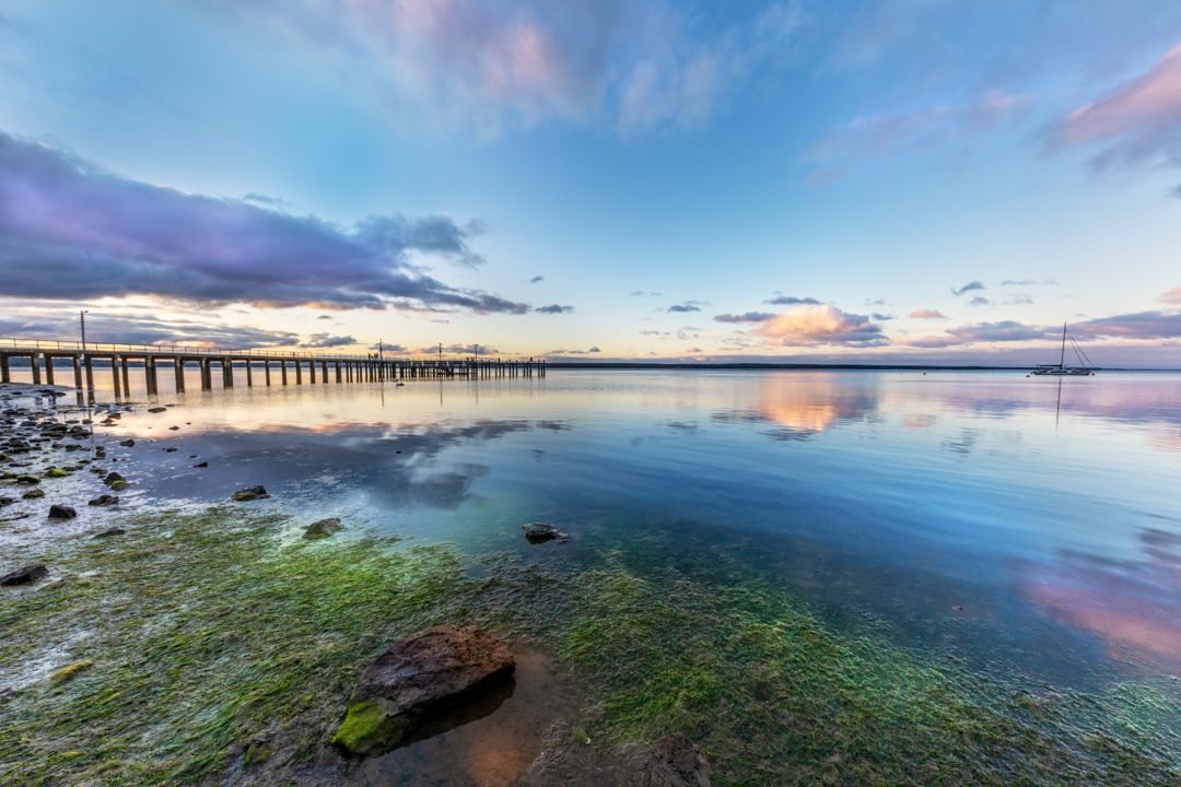 Coastal pier extends over calm water with vibrant sunset colors and algae-covered rocks in the foreground.