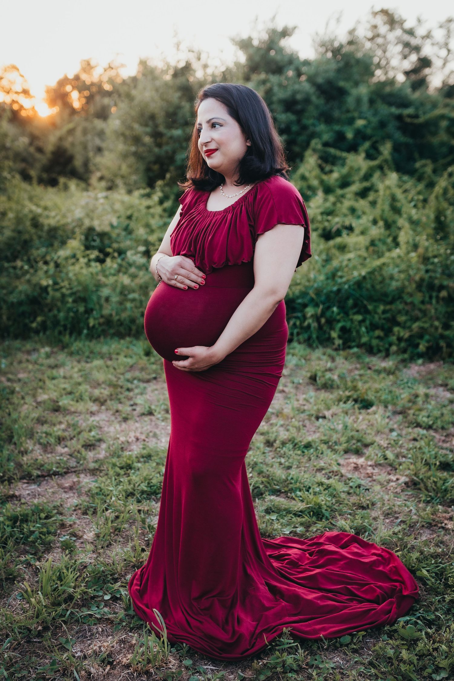Maternity portrait in striking red gown with train against natural foliage.