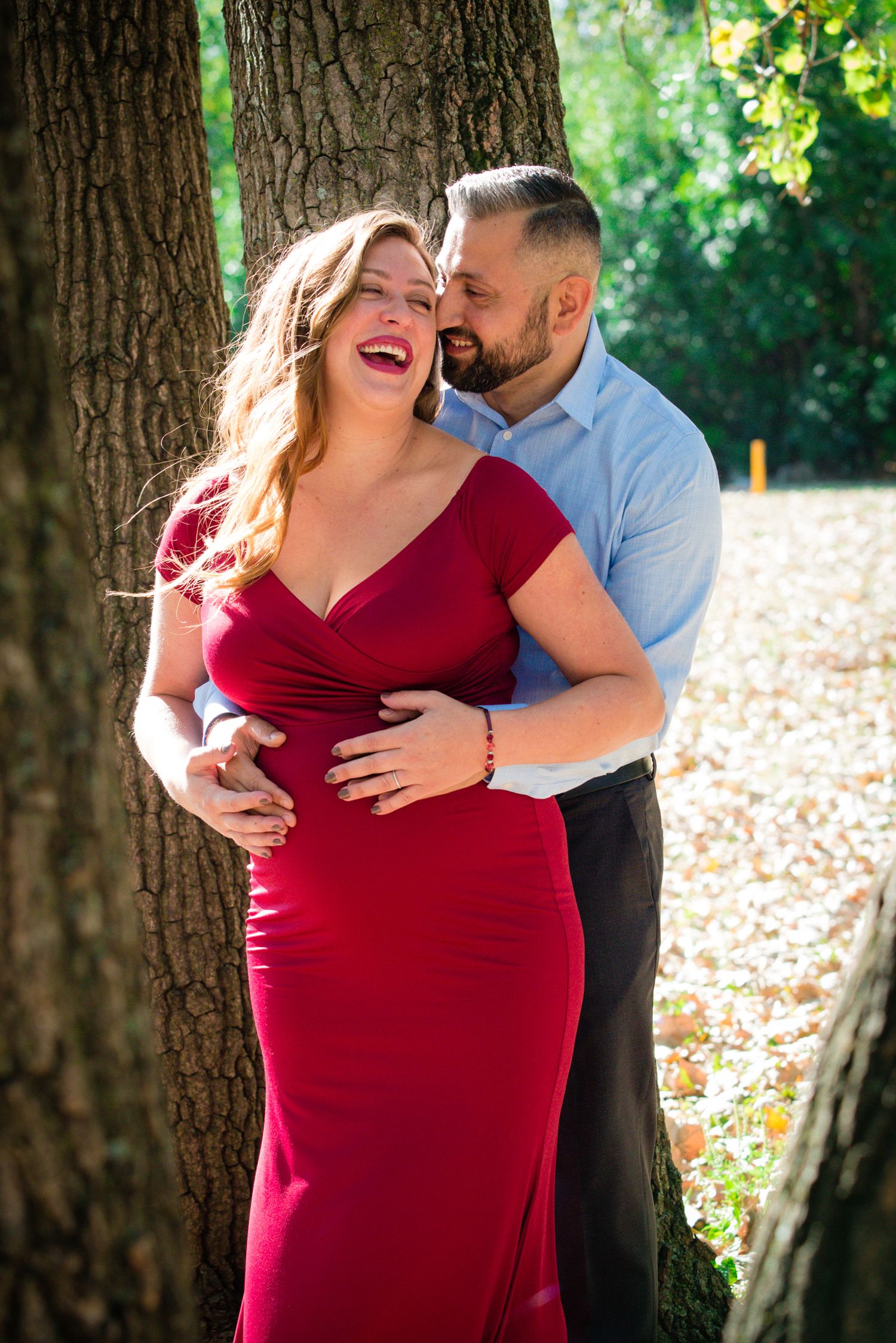 A couple shares a tender moment in front of a large tree trunk while wearing formal attire.
