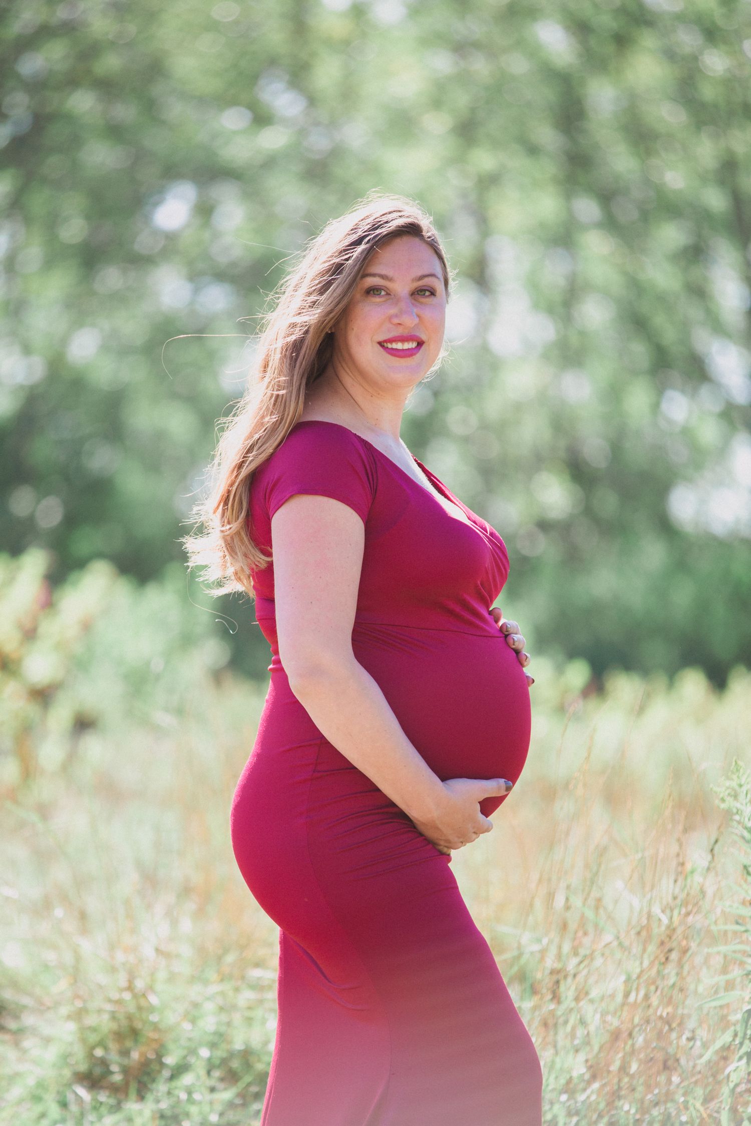 Expectant mother in a red dress poses outdoors with her baby bump in a natural setting.