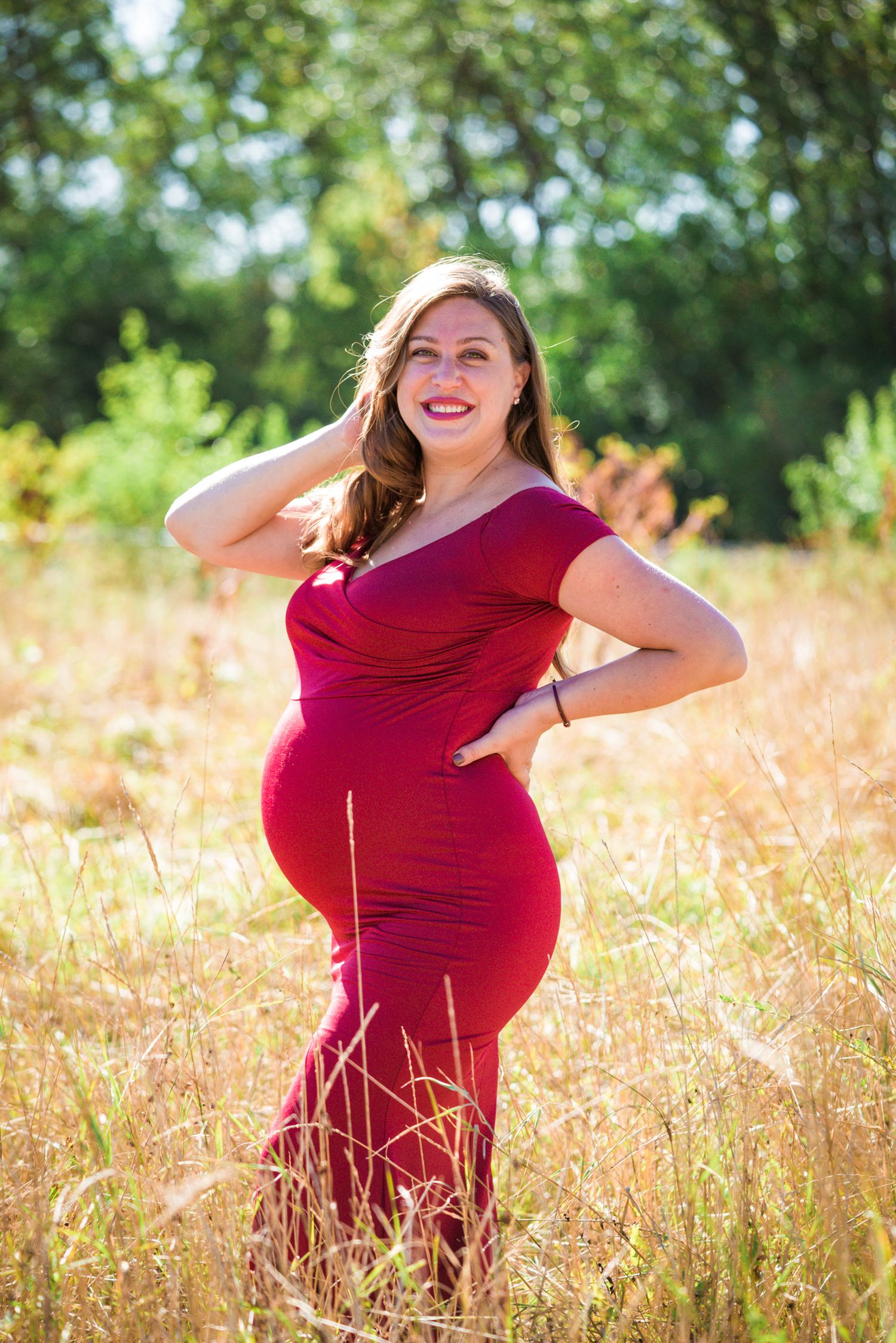 A figure in a crimson maternity dress poses elegantly in a sun-lit field of tall grass.