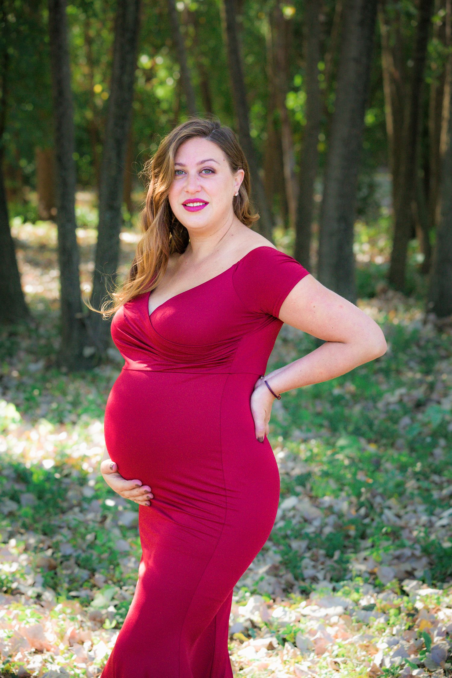 A figure in a fitted red maternity gown poses confidently in a wooded setting.