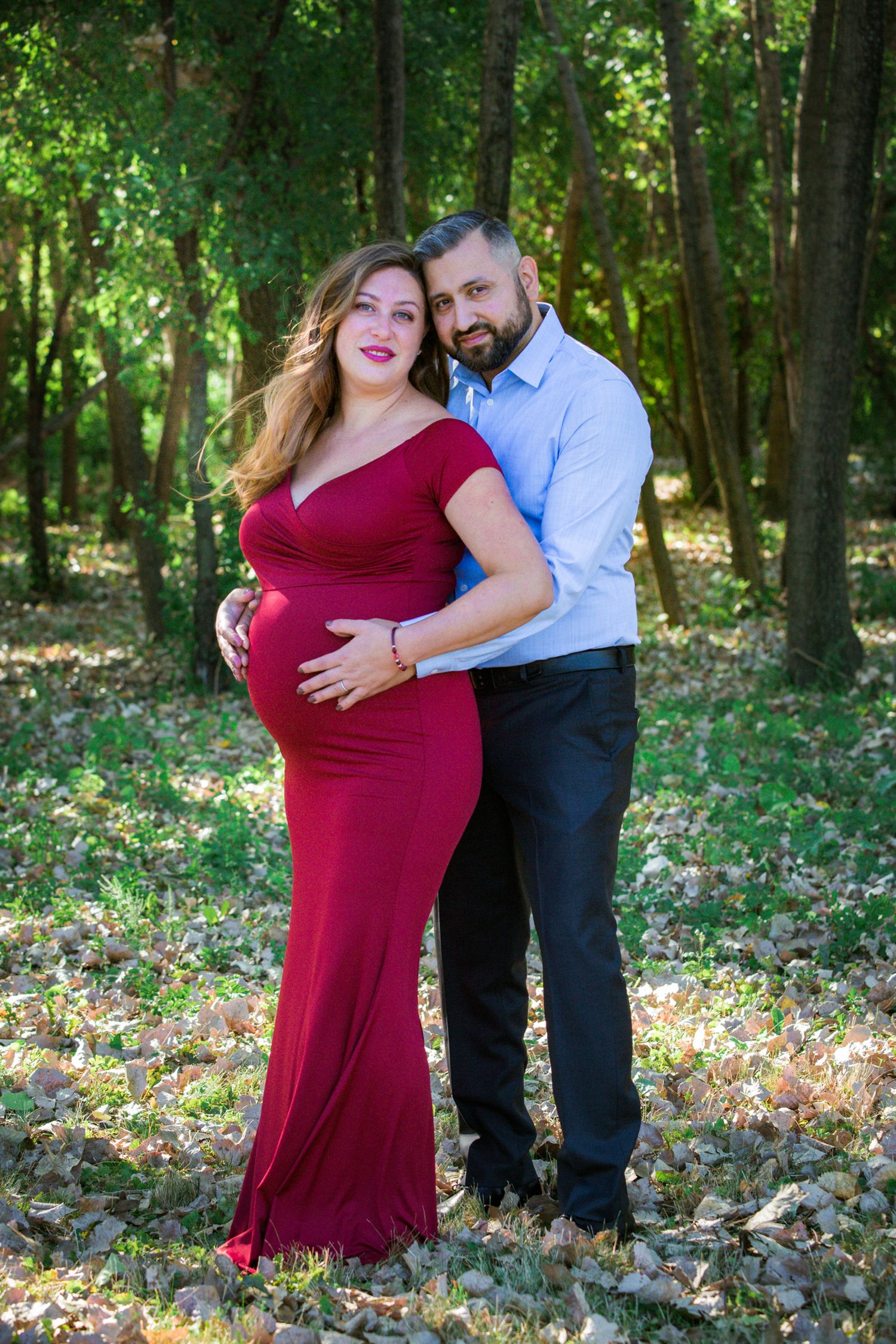 A couple poses for a maternity photo in a scenic wooded area with natural lighting.