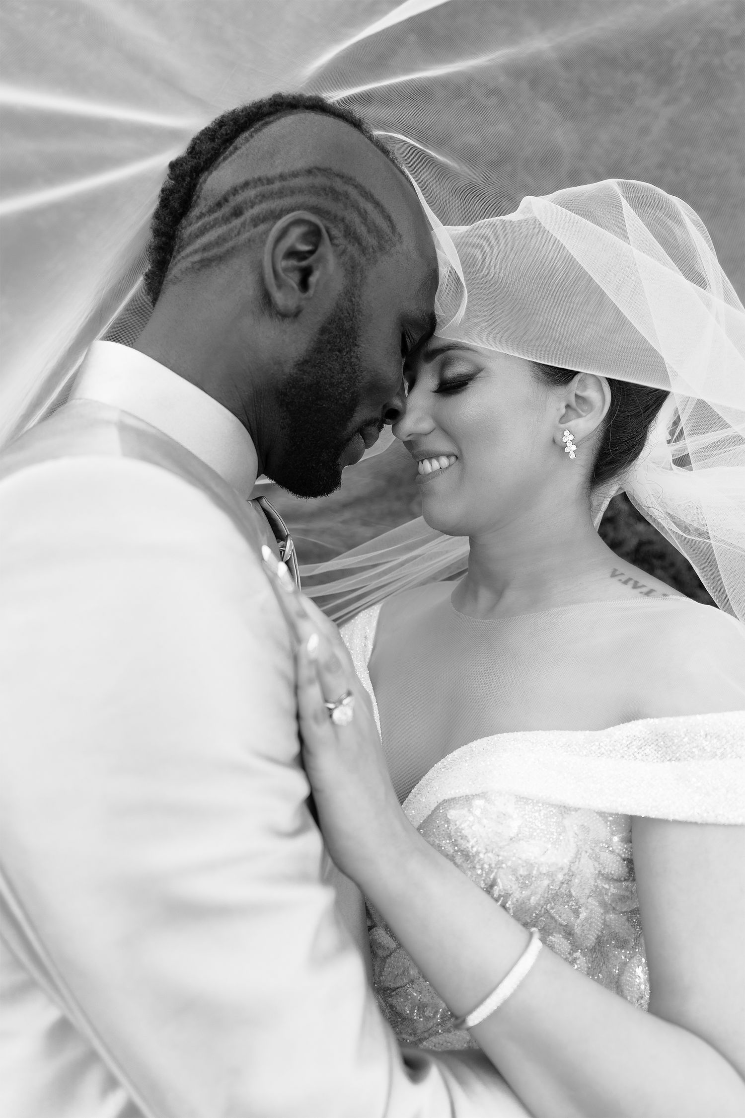 An African man and an Indian woman embrace under her veil just after they were married. White dress, silver tux.
