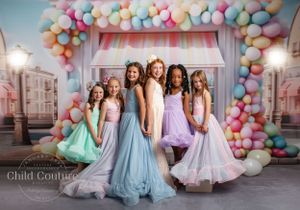 Group portrait with pastel dresses under balloon arch with vintage storefront backdrop.