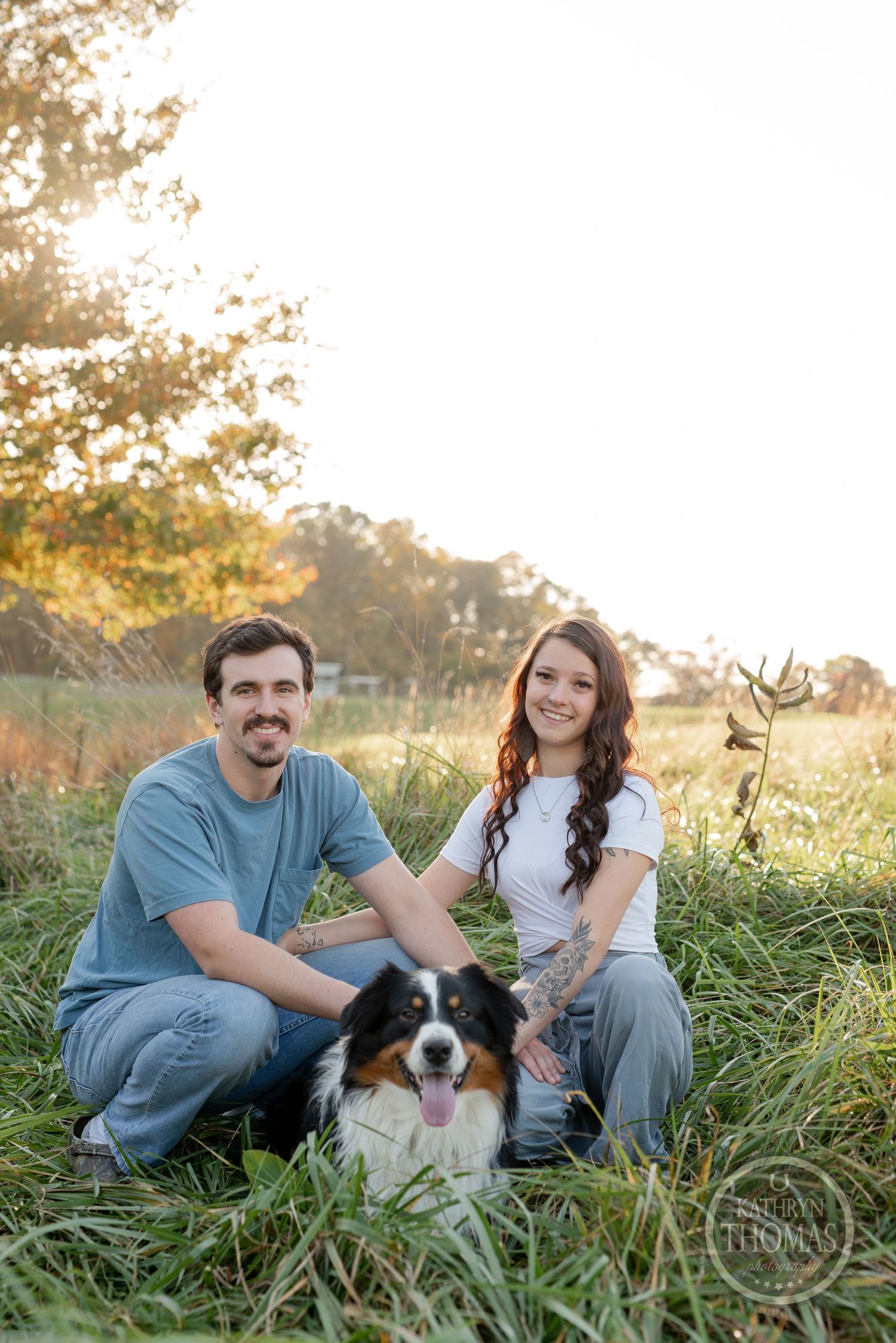 A couple sits with their black and white Border Collie in a sunlit field.