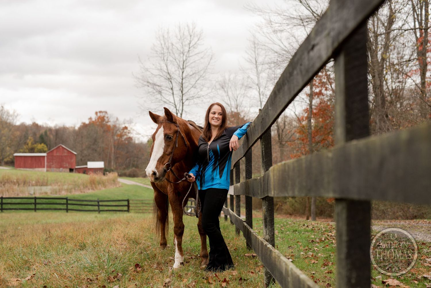 A horse and handler pose alongside wooden fence rails with autumn trees and red barn in distance.