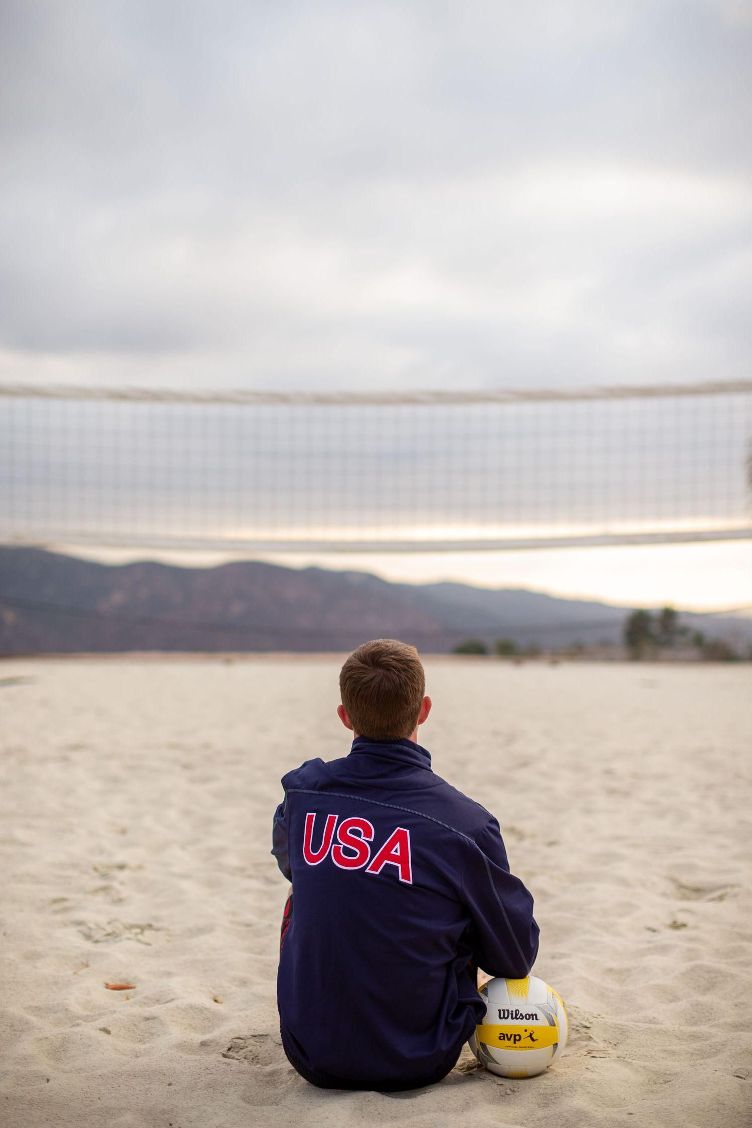 A person wearing a USA jacket sits on a beach with a volleyball, facing mountains in the distance at sunset.