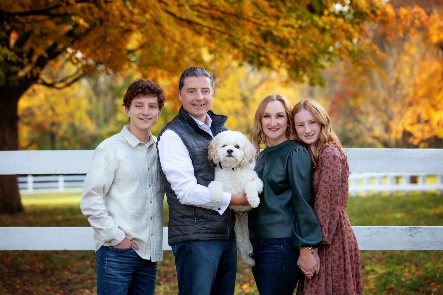 Autumn family portrait with a white dog in front of a white fence surrounded by golden foliage.