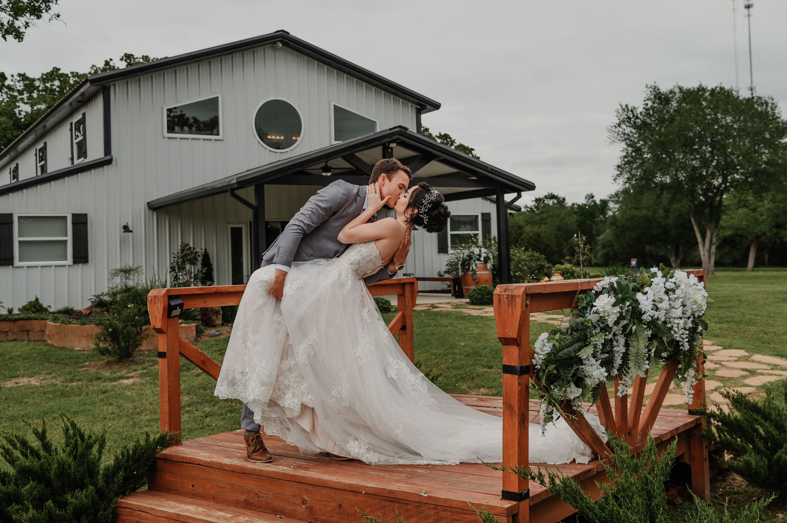 Romantic couple sharing a dip kiss on wooden steps outside a white barn wedding venue.
