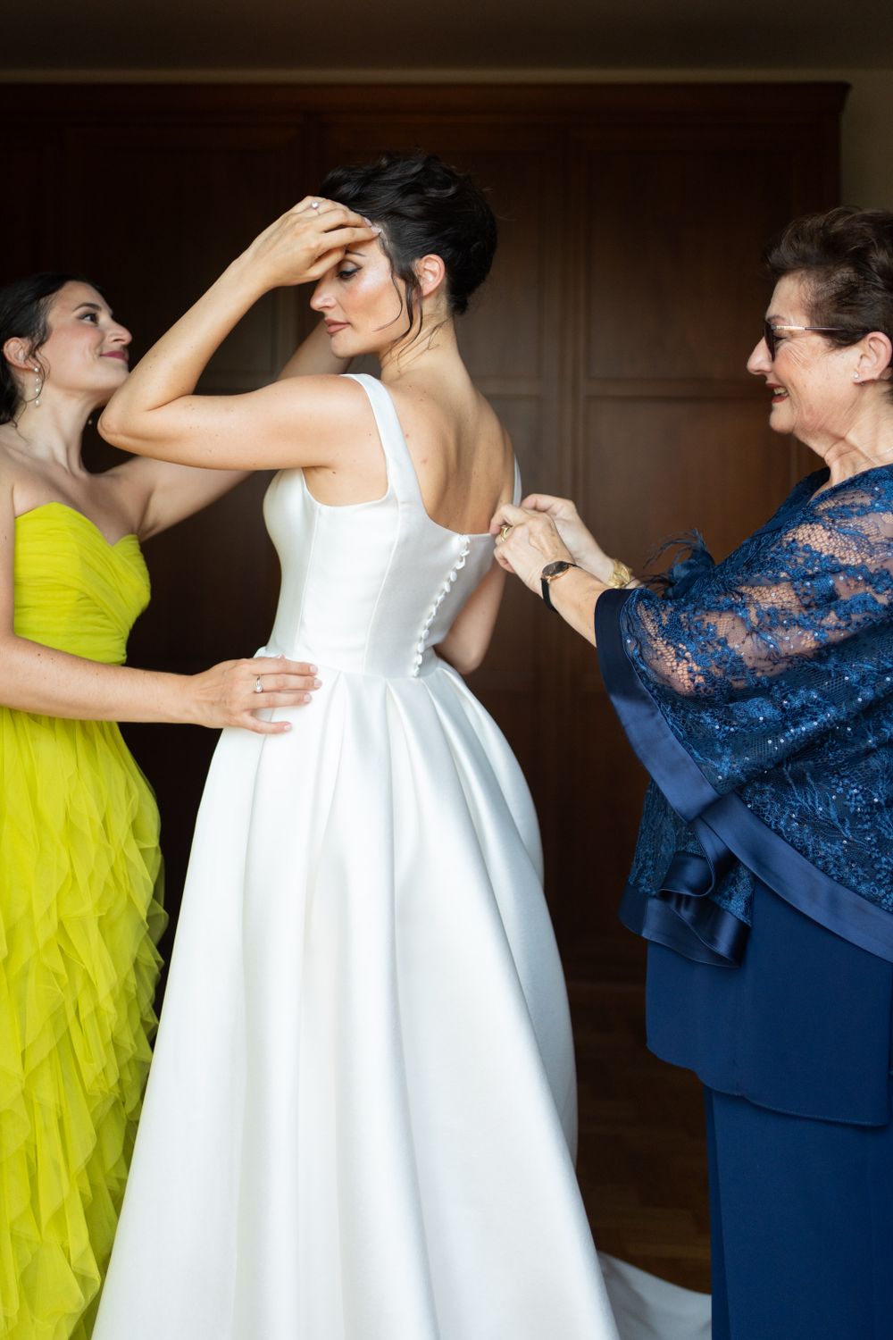Wedding preparation scene shows helpers in navy and yellow dresses assisting with white gown.