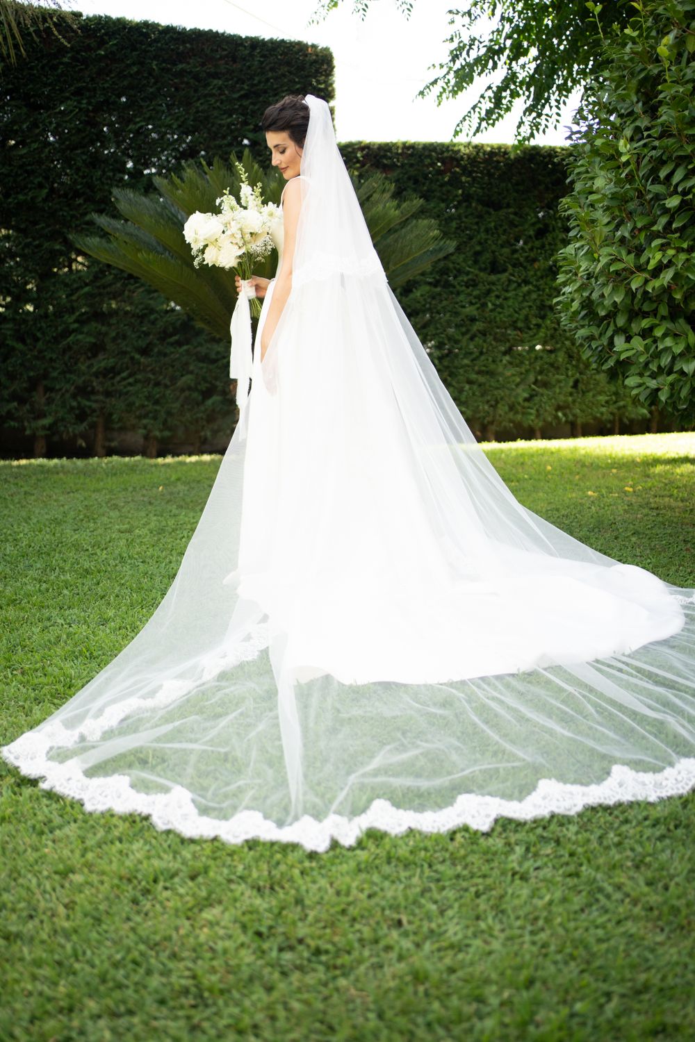 A flowing white wedding dress train spreads elegantly across green grass in an outdoor garden setting.