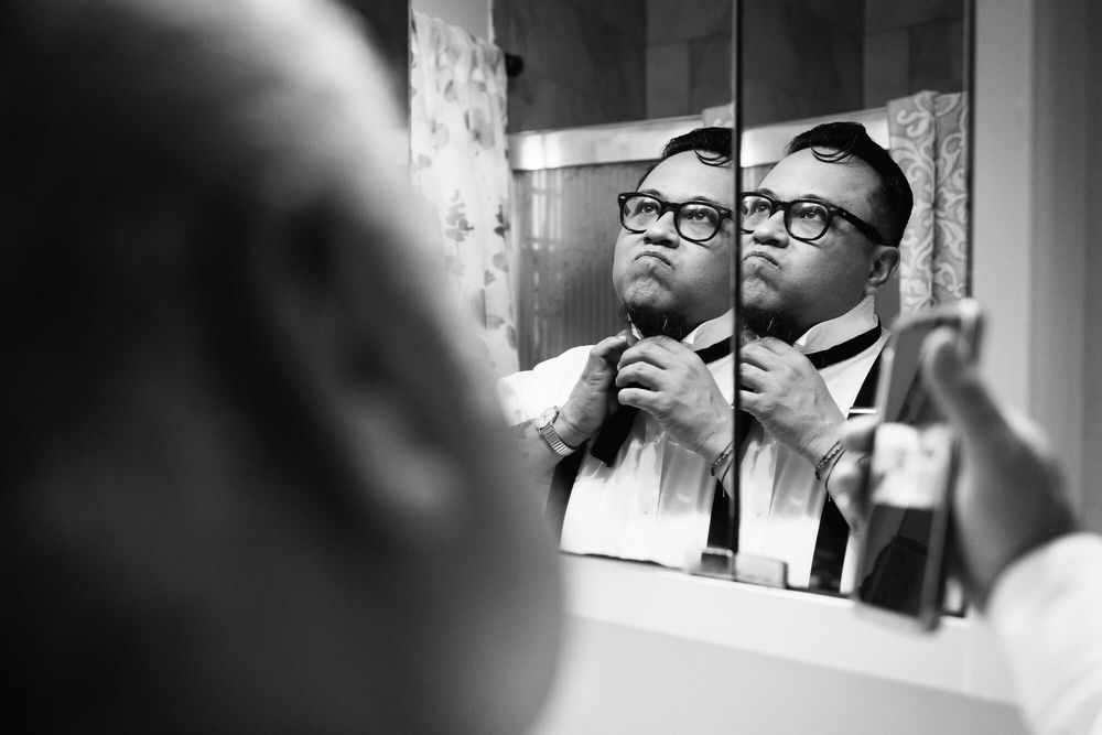A frustrated groom struggles to tie his bowtie while father of the groom holds up phone playing a Youtube video