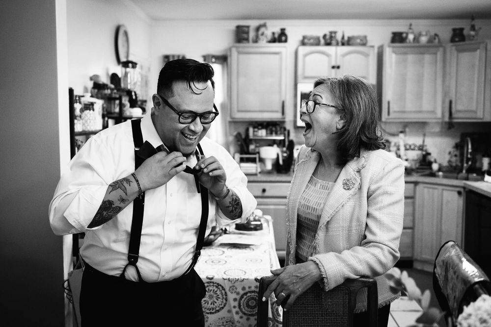 The groom and his mother laugh as he tries to tie his bowtie at their home in Long Island NY.