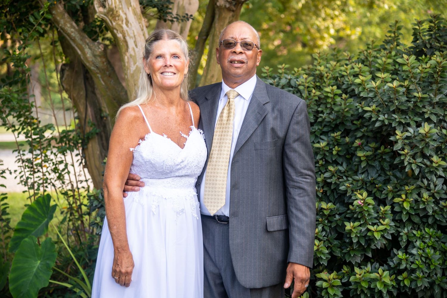 Couple in formal attire poses outdoors in garden setting.