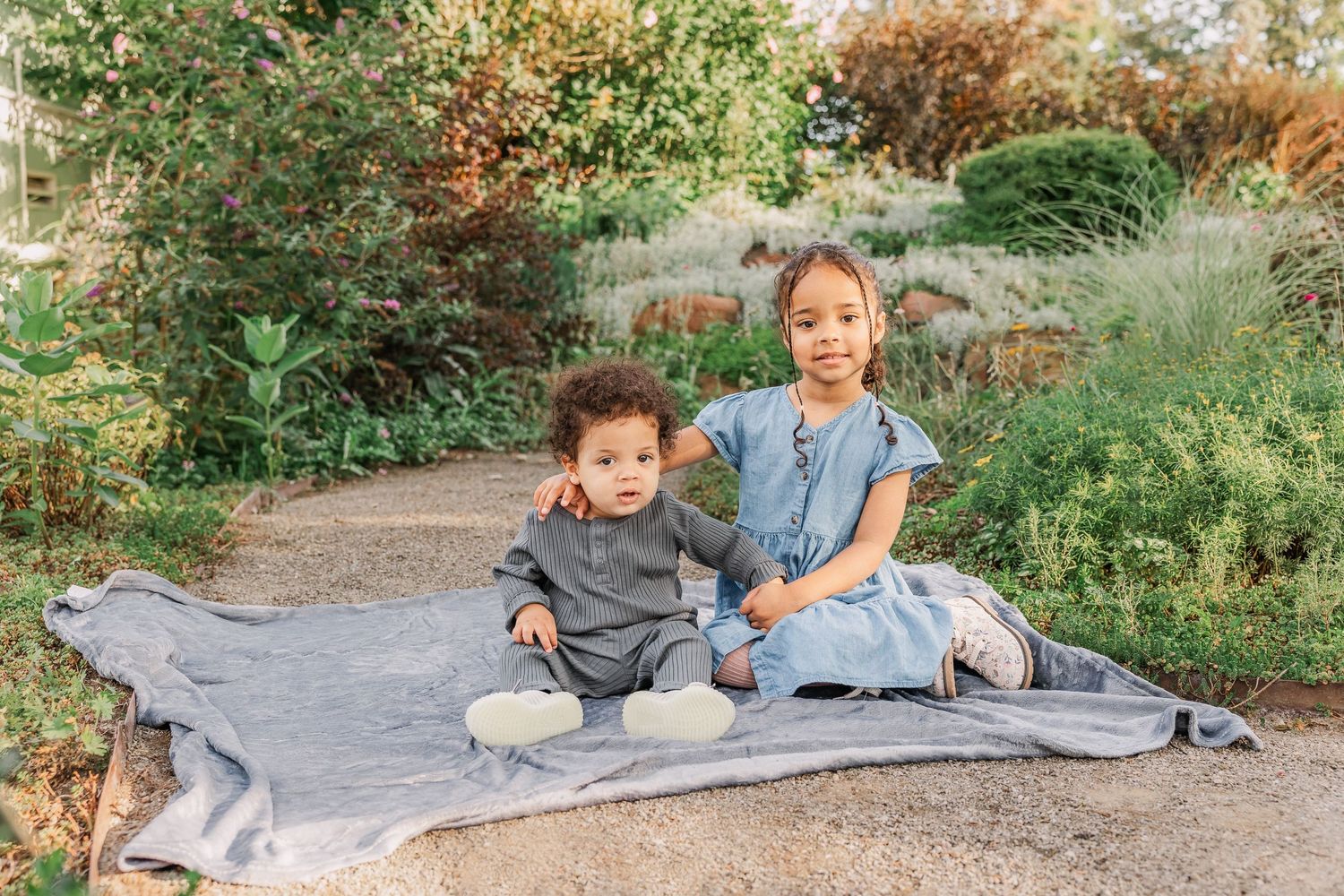 Two children sit together on a blanket in a peaceful garden setting.