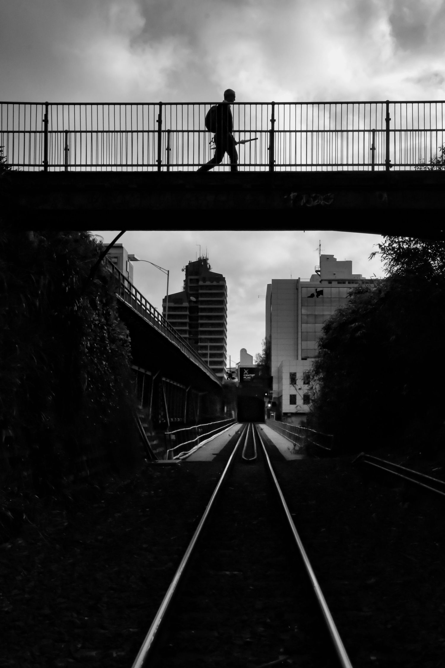 A black-and-white image of train tracks leading into the city, framed by a pedestrian walking on a bridge above.