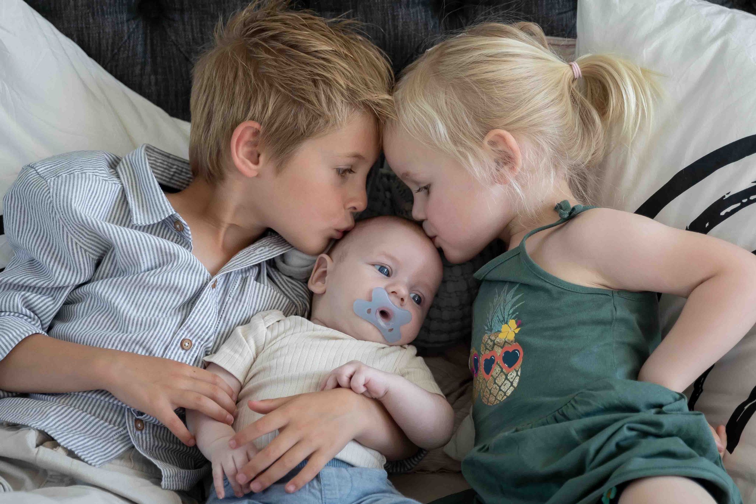 Barbara Kempeneers Fotografie, newborn shoot, grote broer en zus op bed beide geven baby broer een kusje
