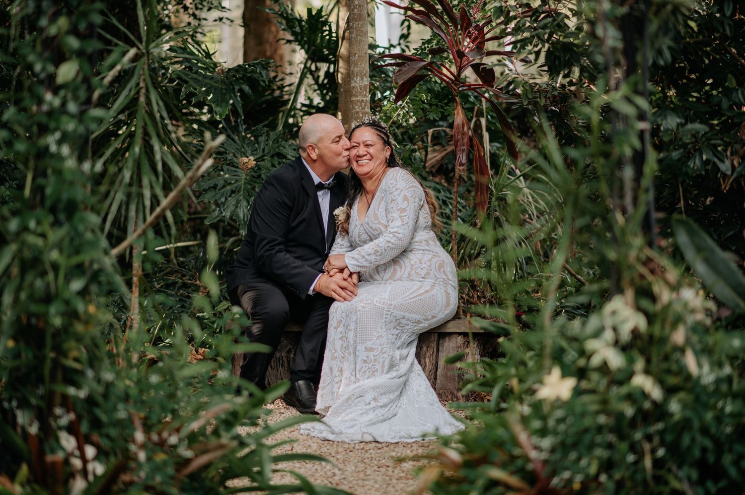 Newlywed couple embraces on garden path surrounded by lush tropical foliage during outdoor wedding photoshoot.
