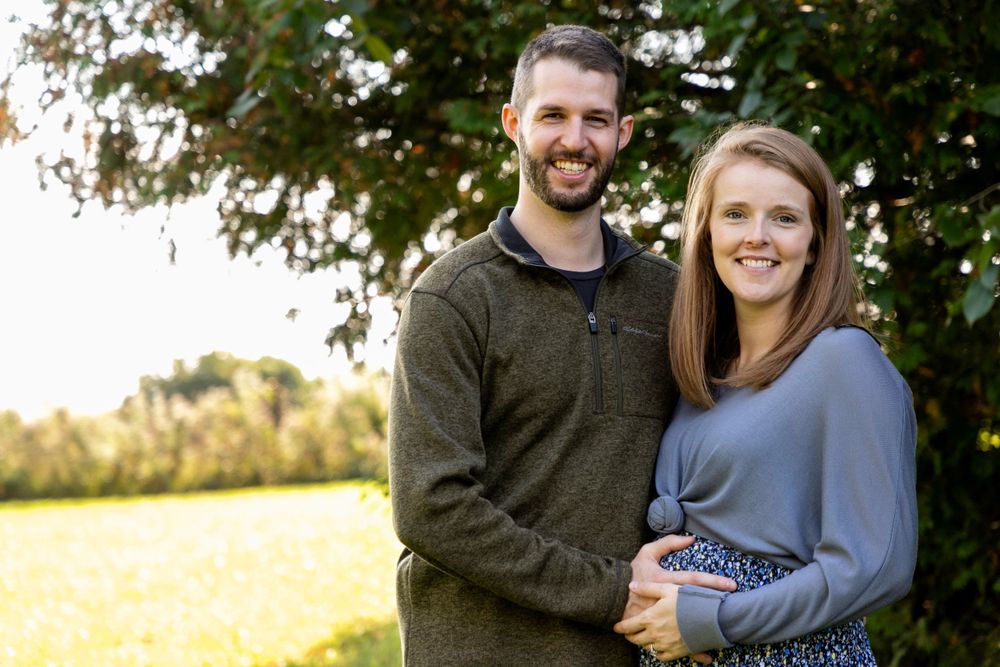 A husband and wife standing next to each other behind some trees and a field holding her pregnant belly