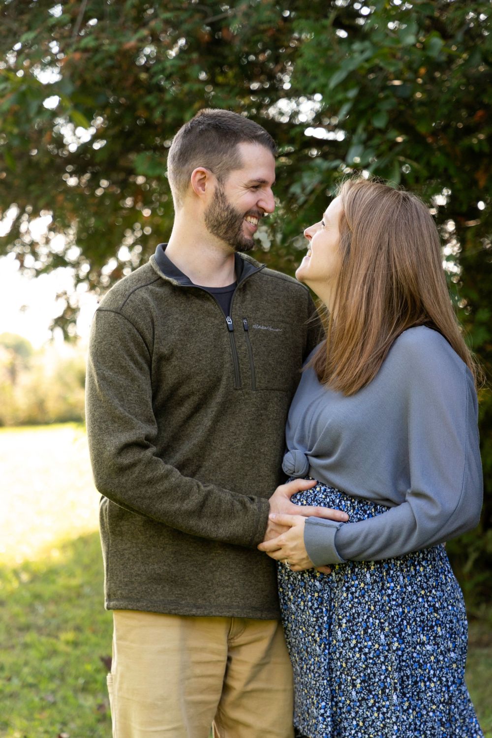 A husband and wife standing next to each other behind some trees and a field holding her pregnant belly