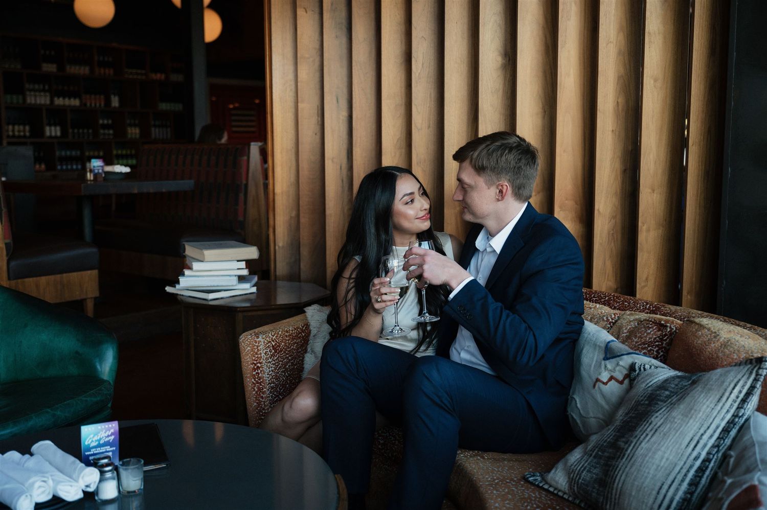 A romantic couple shares wine and conversation on a cozy leather couch in a dimly lit lounge setting.
