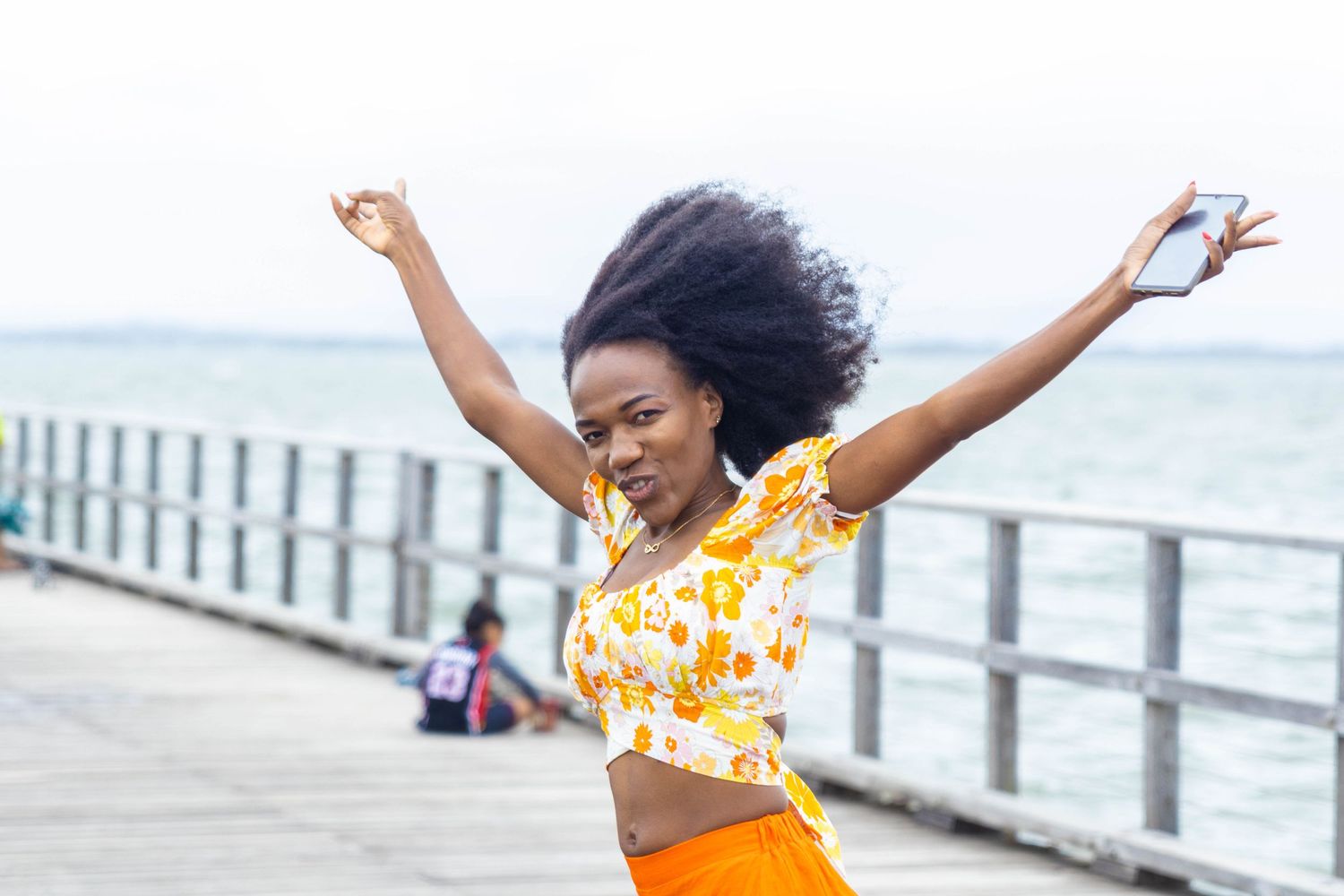 A person in a yellow crop top dances joyfully on a pier by the ocean with arms raised.