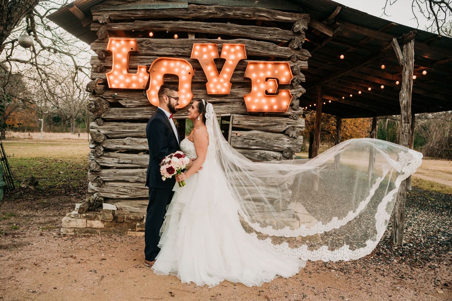 Wedding couple poses outdoors by rustic wood cabin with illuminated LOVE sign and flowing veil in romantic sunset light.