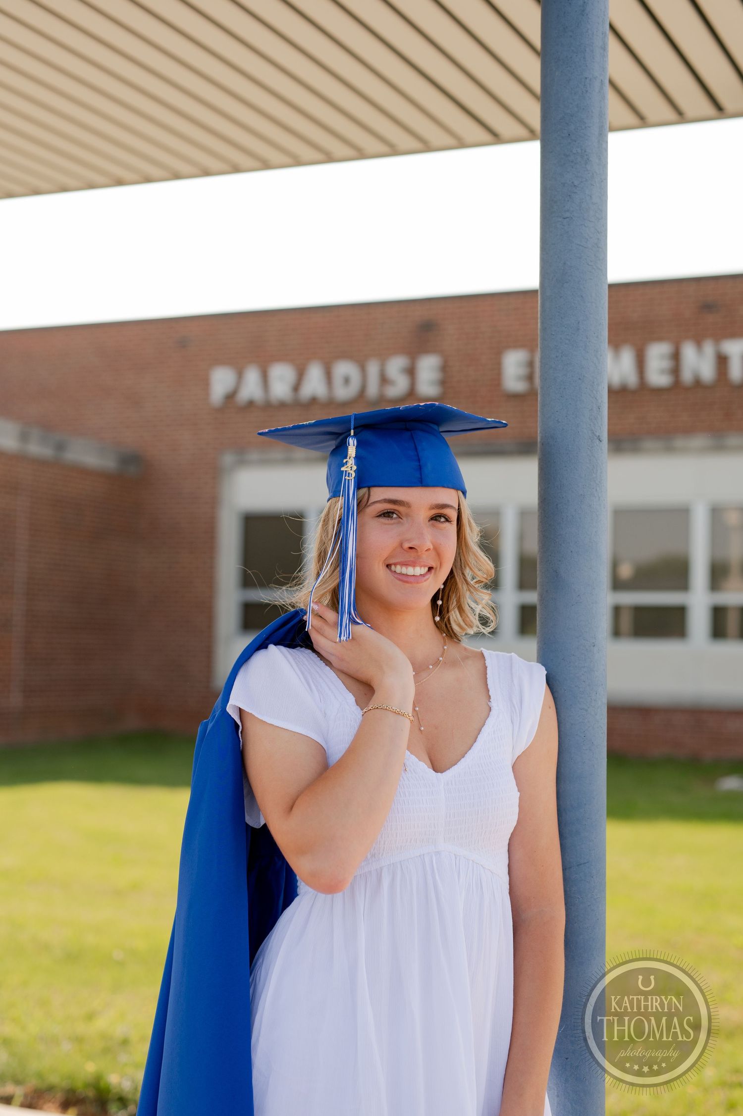 Graduate wearing blue cap and gown smiles while posing at Paradise Elementary entrance.