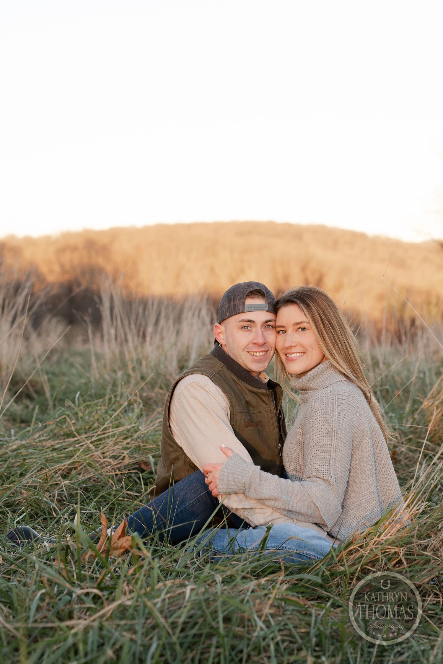 A couple poses intimately in tall grass during sunset with golden hills in the background.