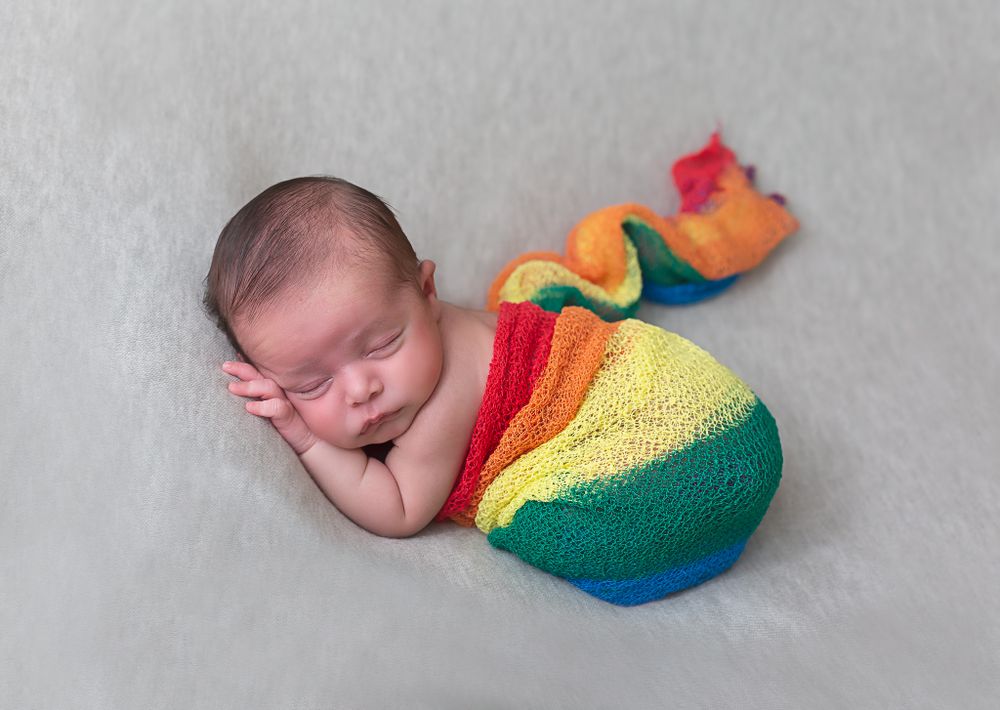 Newborn sleeping in colorful rainbow-themed wrap on gray backdrop.