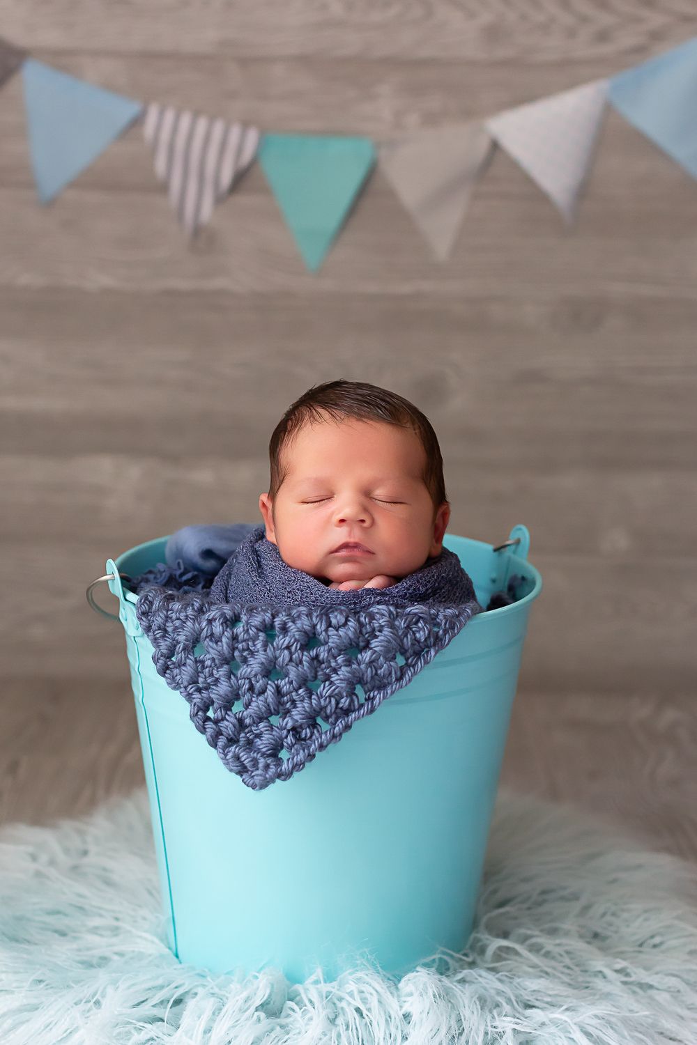 Newborn sleeping in turquoise basket with purple blanket under decorative bunting.