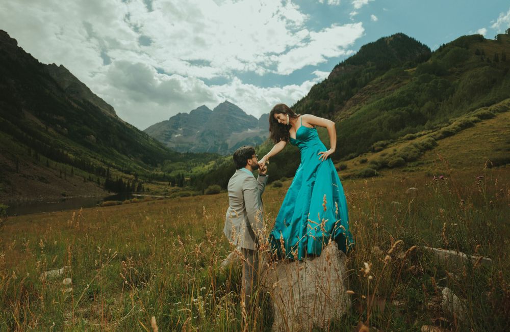 Two figures pose on rocks with a turquoise dress flowing in the mountain breeze against majestic peaks.