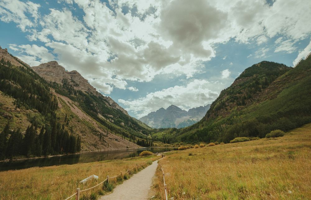 A couple walks along a fenced path through mountain meadows with dramatic peaks in the distance.