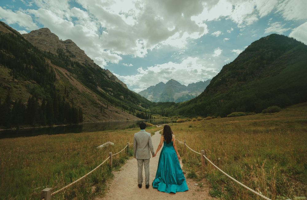 A couple walks hand in hand down a mountain path with a flowing turquoise dress catching the light.
