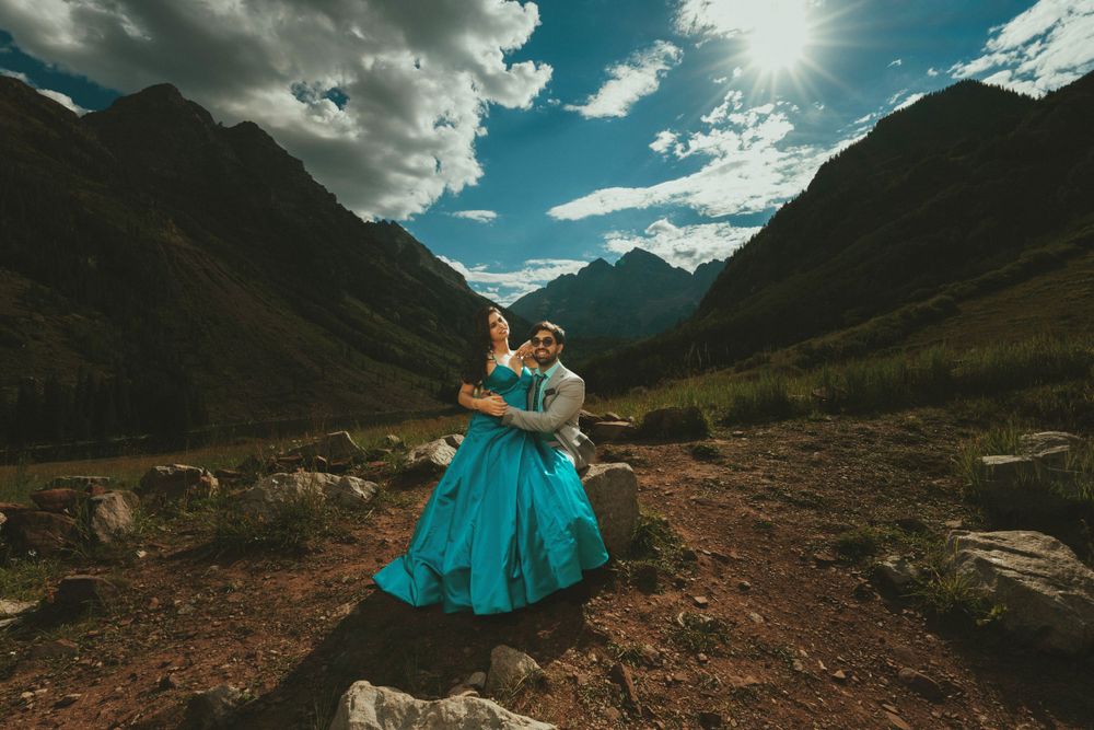 A turquoise formal dress stands out against dramatic mountain scenery with sunburst through clouds.