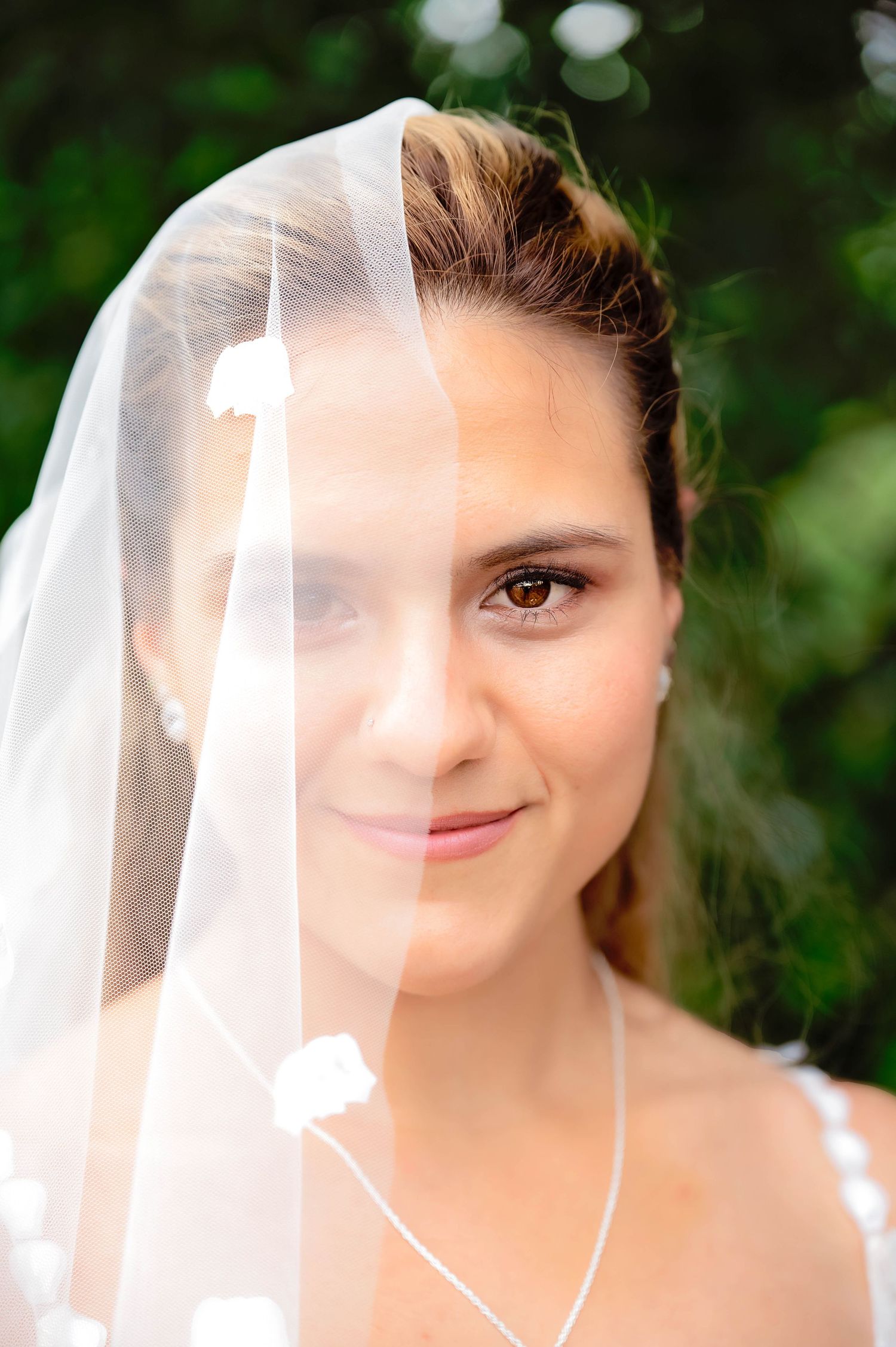 Close-up portrait of a bride wearing a sheer white veil against a blurred green outdoor background.