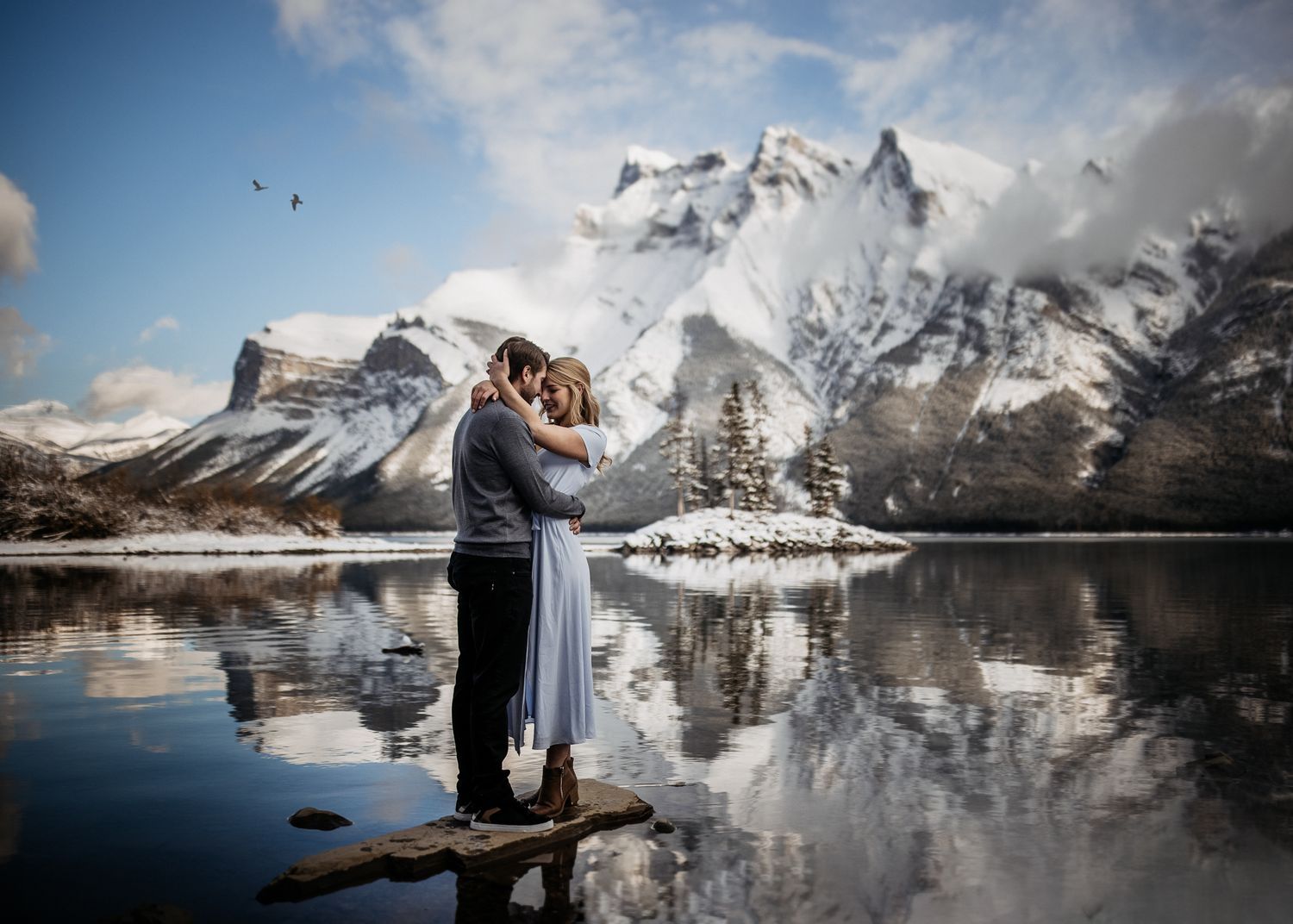 Dramatic winter wedding photo at mountain lake with snowy peaks and reflections.