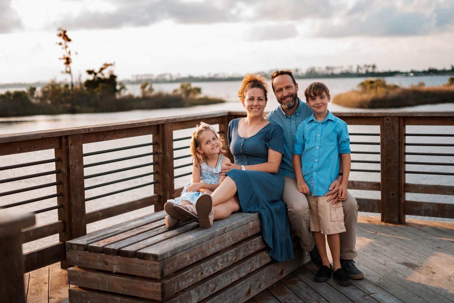Happy family of four posing on a wooden waterfront deck at golden hour with scenic water views.