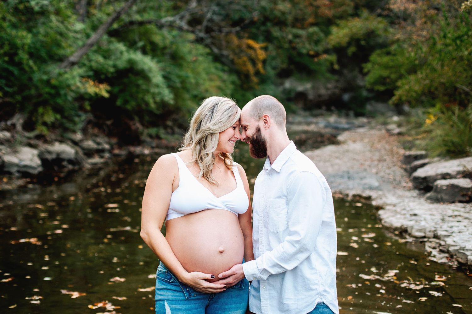 expecting mom wearing jeans and white sports bra poses with spouse near a creek in lee's summit missouri