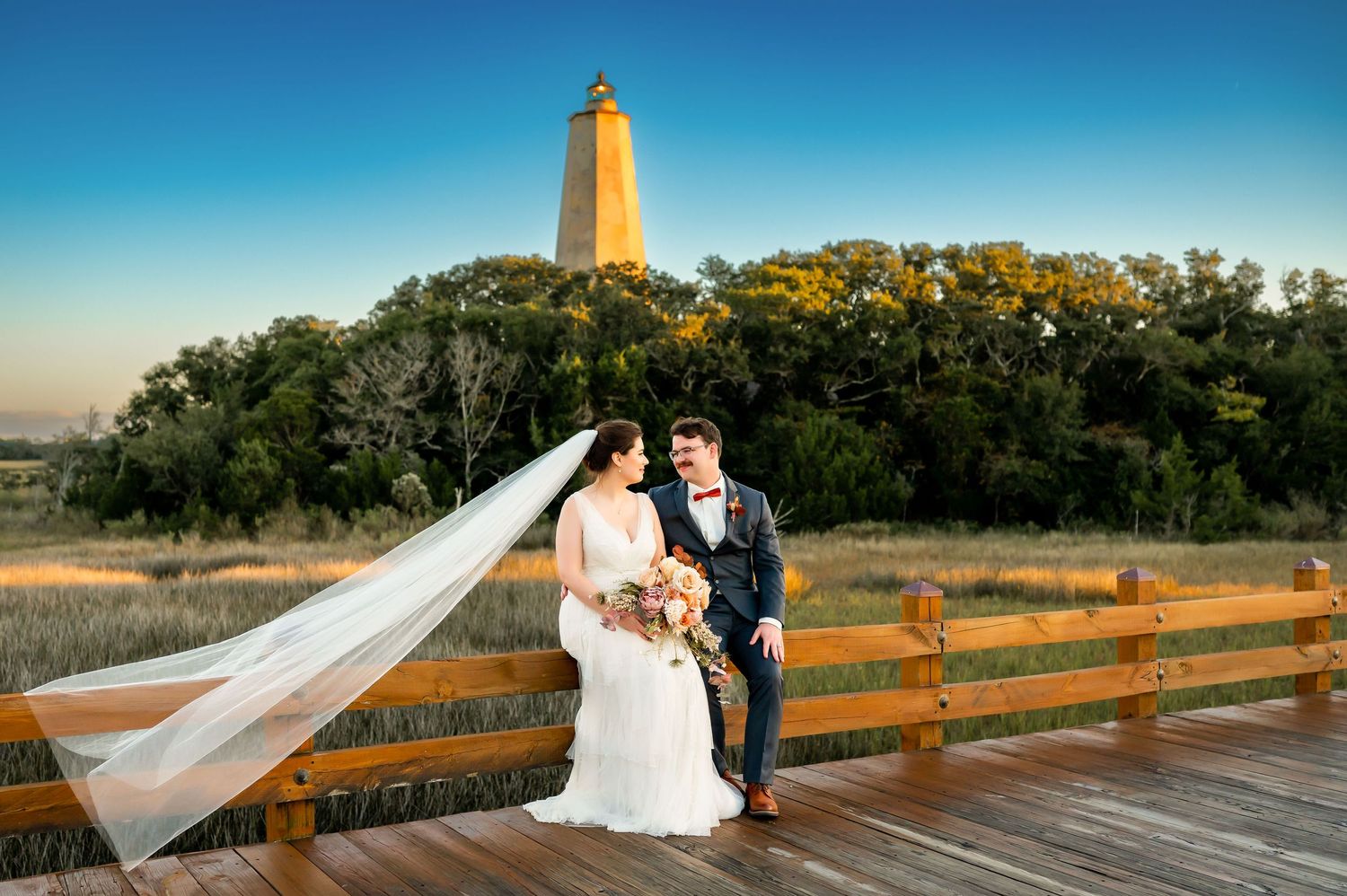A romantic wedding couple poses on a wooden deck with a flowing veil against a natural outdoor backdrop.