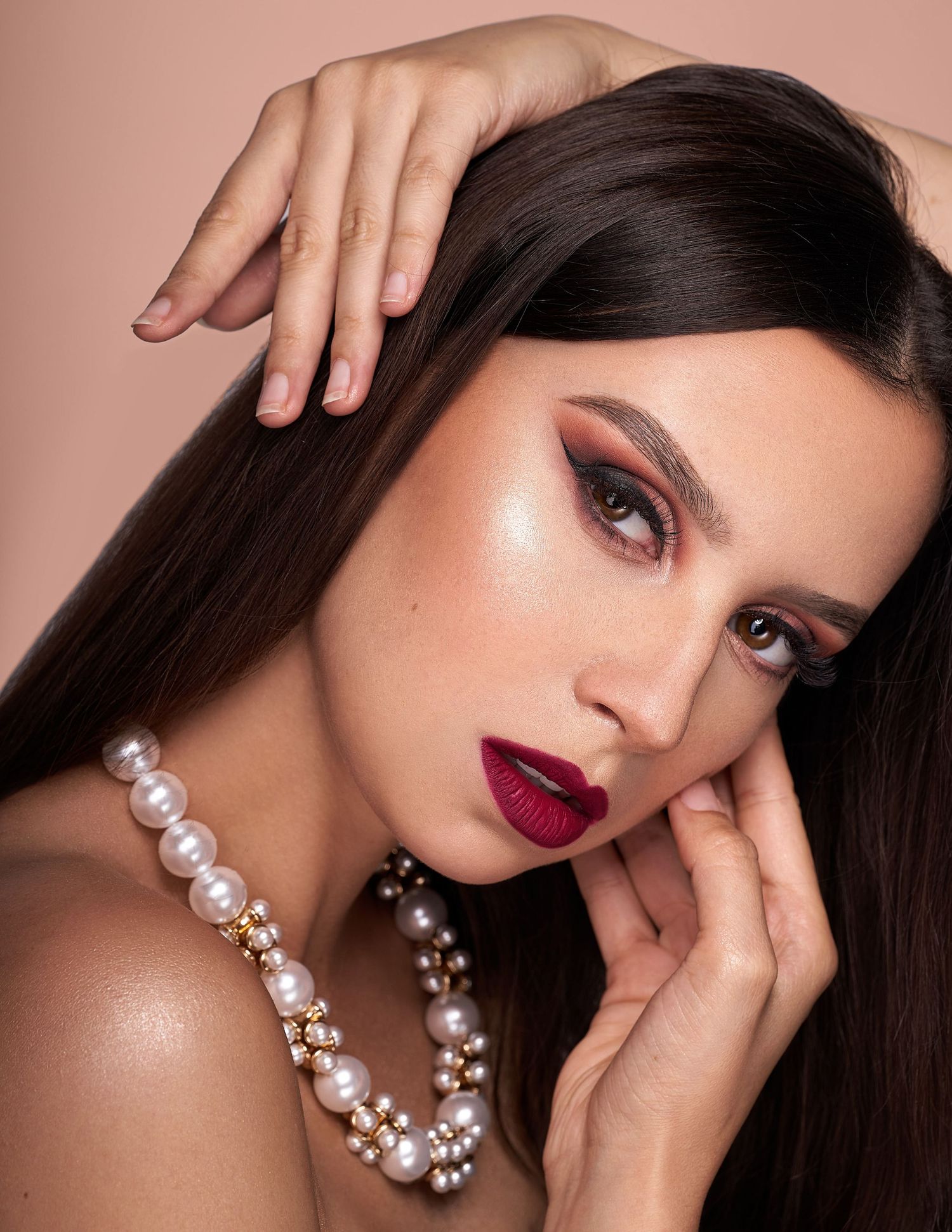 Striking glamour portrait featuring dark hair, bold red lipstick, and an elegant pearl necklace against a pink background.