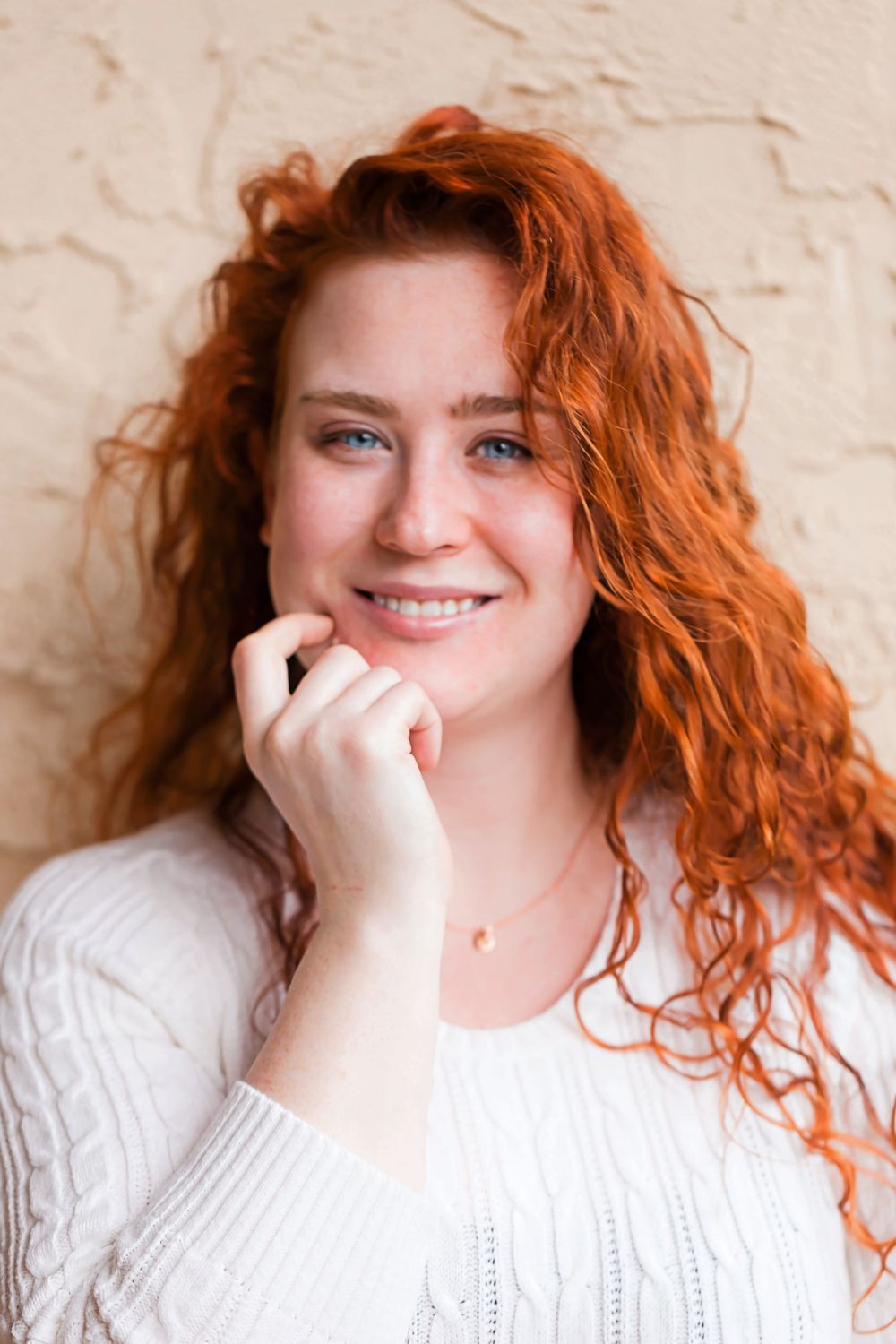 Young woman with curly red hair and freckles smiling warmly in a white sweater.