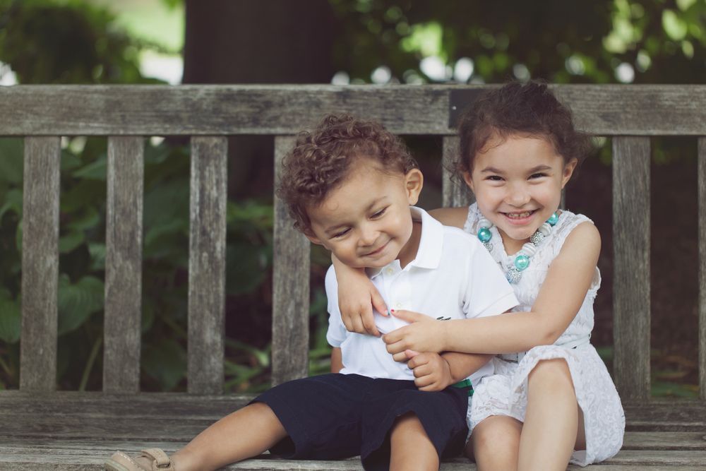 Two young children hugging and smiling on a wooden bench outdoors.